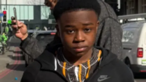 Abdul is pictured wearing a black hoodie and earphones looks towards the camera on a city street, with a car and a cyclist visible in the background.