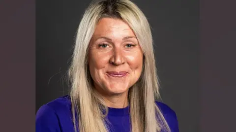 Peterborough Women's Aid Mandy Geraghty. She has long blonde hair and is wearing a bright blue top. She is smiling towards the camera