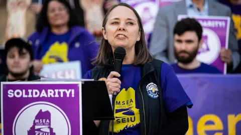 Getty Images Woman in blue and yellow t-shirt holds black microphone under chin while speaking behind a purple sign reading SOLIDARITY! before a small group of people also dressed in blue and yellow and holding signs