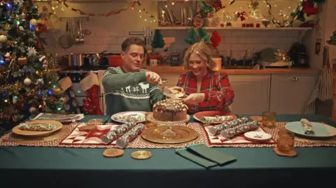 Waitrose Matthew Horne and Joanna Page sat at a Christmas decorated kitchen table. Horne is cutting a piece of cake for Page. Both are wearing festive outfits, with Horne wearing a green Christmas jumper and Page wearing red tartan pyjamas.