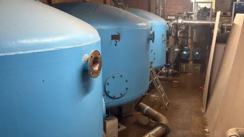 Matt Day Three blue cylinder tanks are lined up next to each other on the left. There are multiple pipes coming from them and there is wood lined up alongside the right. 