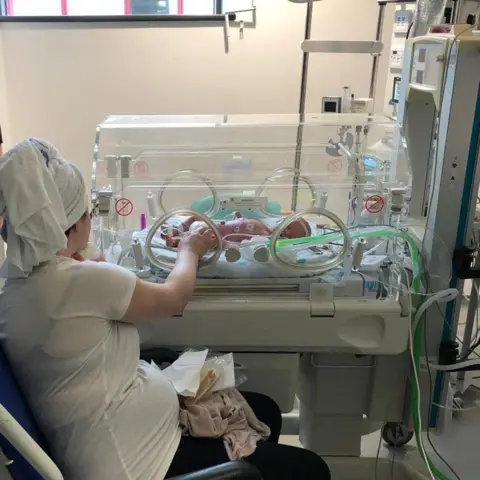 Kianty and Conor Simpson Kianty with her hair wrapped up in a towel sits in a hospital wheelchair in front of an incubator. She's reaching through a circular window in the incubator to touch her baby son's hand.