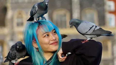 Summer Loh, a young woman with blue and pink hair who is standing on top of Norwich Markets in the city. In the picture she has a pigeon on her head, shoulder and elbow.