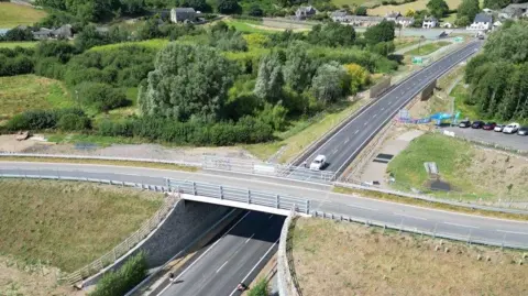 Aerial view of a road crossed by a road bridge. It is newly built, as can be seen by the freshly grassed banks to either side. The setting is rural with a patch of trees to the left and a small scattering of houses in the distance.