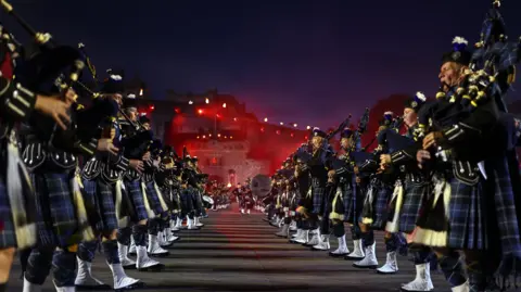 Getty Images Two rows of pipers during a preview of the Royal Edinburgh Military Tattoo at Edinburgh Castle, which can be seen in the background shrouded in red light.