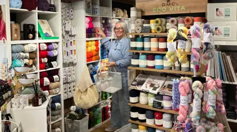A woman is selecting yarns of wool from a wall of shelves filled with different coloured wool