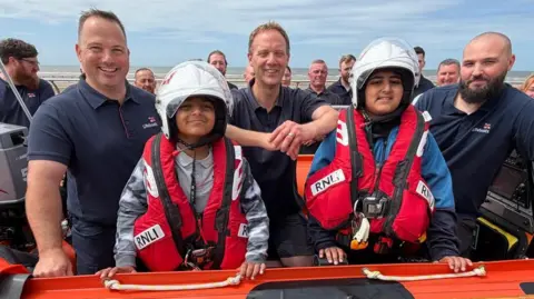 Yunus Mulla/BBC Both young boys have light grey helmets and red life-jackets and are flanked by RNLI members.