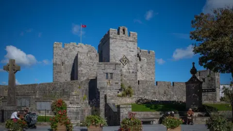 MANX SCENES A medieval castle which had a square tower with a clock face on it and a Manx flag flying from the battlements with blue sky behind.
