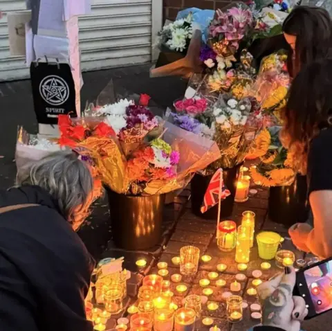 Three women kneel down next to a number of lit candles on the ground. Behind the candles are a number of bunches of flowers in varying colours.