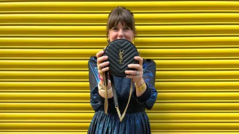 Caroline Jones Caroline Jones is wearing a shiny blue long sleeved dress and is holding a round, black YSL bag with a gold chain strap in front of her. Caroline has dark brown hair which is tied up so that you can only see the fringe.