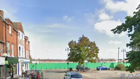 Google A plot of land behind green fencing occupies the centre of the picture. There are three-storey terrace houses and shops to the left, with cars parked outside. There is a tree to the right and another in the centre, in front of the fencing.