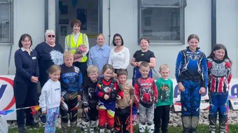 LITTLE KIPPERS Ten children, some of whom wearing colourful motorbike gear, smile for the cameras. Five adults stand behind them in front of a grey hut.