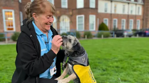 Yvonne Batten crouches down to feed a treat to Hugo, a border terrier, who is standing on his hind legs with his front paws on her knee. Yvonne is wearing a black hoodie and light blue polo shirt, the PAT volunteers uniform. Hugo is wearing a yellow bib.