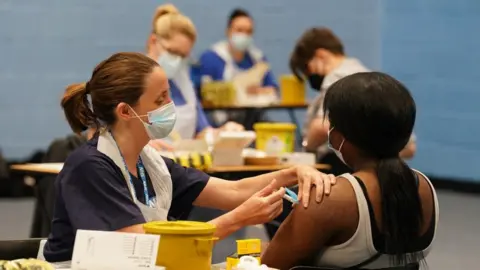 PA Media A nurse administers a vaccine does to a girl who is seated and faces away from the camera. The rest of the image is blurred but others can be seen also receiving their dose