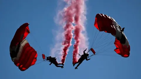 Getty Images Two people in the sky with red parachutes. They are close to each other, with a red trail of smoke coming from their feet.