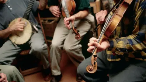Getty Images Musicians in a circle, playing instruments including a banjo and fiddles.