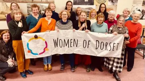 Women of Dorset A group of 17 women posing for the camera. The people in the front of the group are holding up a long white banner saying Women of Dorset.