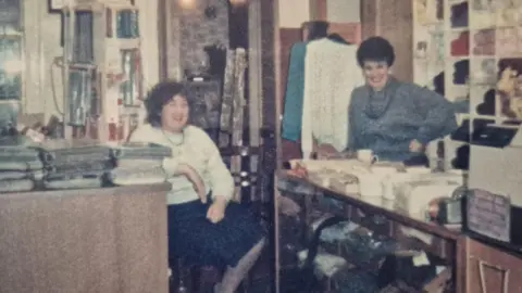 Christine's Wool Shop Two women with short dark hair are wearing jumpers and standing inside a shop with piles of books on tables and yarn stacked in the background. 