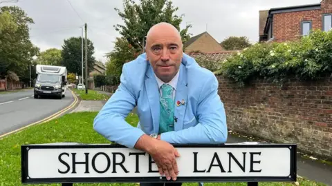 A man in a pale blue suit leaning on a black and white street sign which reads Shorter lane