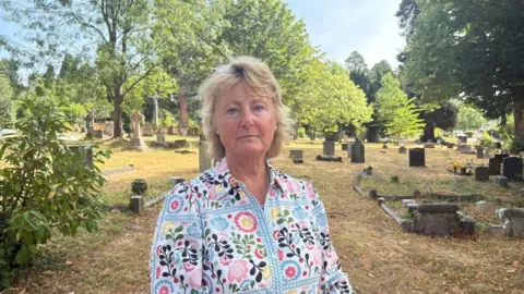 A woman with short blond hair and a patterned pale blue dress is standing in a cemetary on a sunny day.