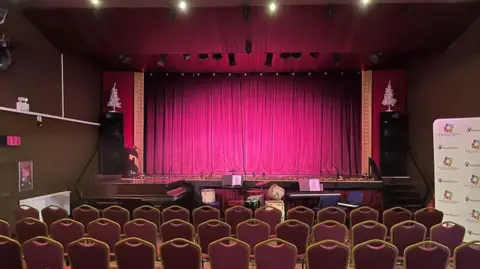 O interior do teatro. Há um palco repleto de cortina vermelha em frente ao qual estão sentados