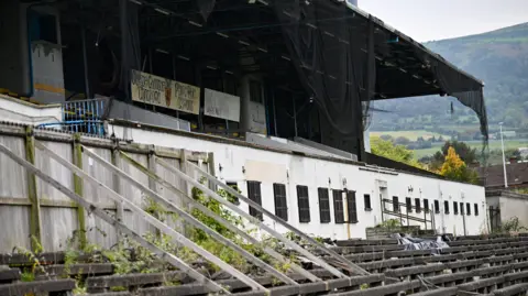 Getty Images A rundown stadium. Planks of wood are attached to a fence to keep it erect. Netting is hanging from the roof. Plants are overgrown.