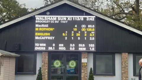 An electronic scoreboard attached to the side of a black building. It is displaying the score of a cricket match. 