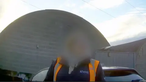 A woman with a blurred face wearing an orange high-vis gilet and a blue fleece. There is the arc of the roof of a building in the background.