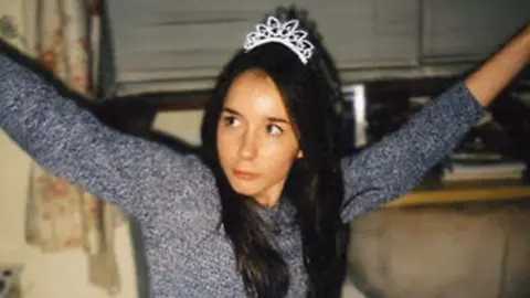 A picture of Fiona Braidwood wearing a long sleeved grey top and a sparkly tiara on her long dark hair. She is sitting in a living room in front of a window, lifting both her arms high and looking off to the left of the camera.