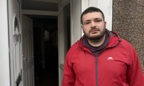 A bearded man wearing a red waterproof jacket stands in the doorway of a house, looking at the camera, with a dim hallway visible behind him.