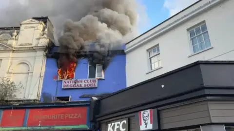 BBC Flames and smoke pour out of the upstairs windows of Nations Club on Union Street. The building is blue and is above a pizza shop. In the bottom of the image a KFC logo can be seen, which is the building next door.