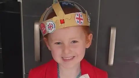Family hand out Lila is smiling directly at the camera. She is a gold, handmade crown which has stickers on it. She has a blue beaded necklace and a red jacket.