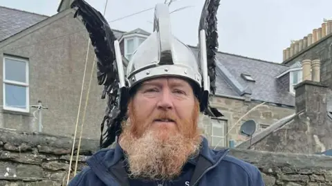 58-year-old Lynden Nicolson is wearing a black-feathered Viking helmet. A ginger-gray beard covers his face and he is wearing a dark-blue jacket with a writing that reads "Scottish Water".