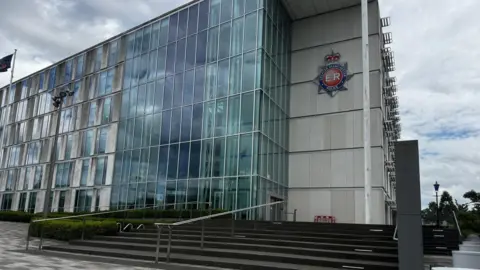 An external view of a multi-storey glass-fronted office building with a Greater Manchester Police crest mounted onto a concrete recess. 