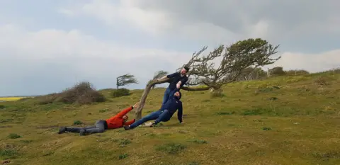 Veronika Voitkevica Three people pretend to cling to a tree in high winds in Newhaven Beach