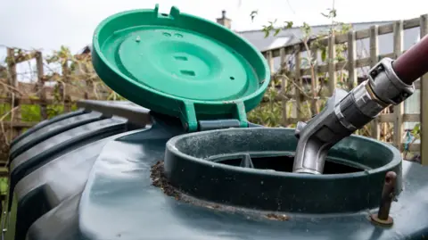 A large plastic heating oil tank in a garden with its top open and a hose filling it.