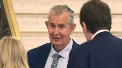 A man with short grey hair, wearing a navy suit, blue shirt and silver tie. He is speaking to two people, who are facing with their backs to the camera, also in formal wear. They are stood beside an ornate stone staircase at Stormont.