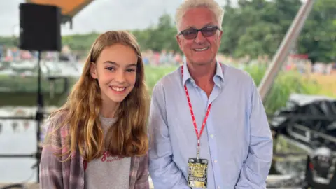 Jon Wright/BBC Anna, an 11 year old girl, stands next to Melvin Benn on the waterfront stage with riverbank and staging behind