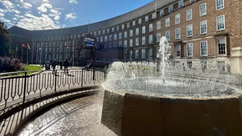 BBC A wide shot of the front of City Hall in Bristol on a sunny day with fountains shooting water up in the foreground