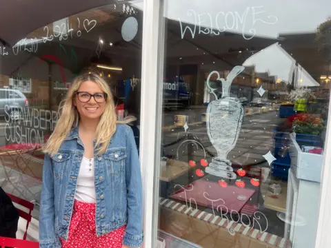 A smiling woman stands beside her decorated shop window that reads "welcome to troon" around a drawn Claret Jug trophy.