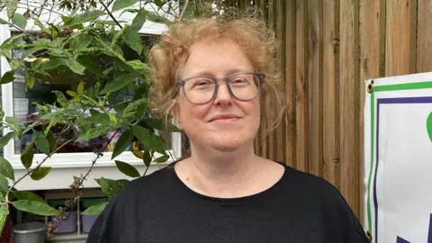 Jack Maclean/BBC Woman with blond hair and wearing glasses stands outdoors next to a fence with a poster (out of view). There is shrubbery behind her.