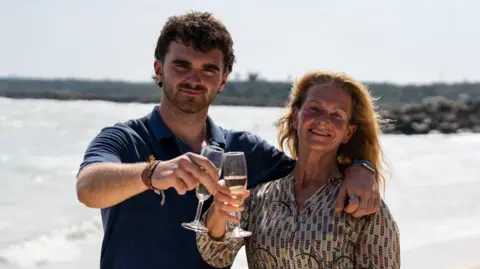 PA Media Tom and Caroline Bridge standing outside on a beach both holding a glass. Tom is on the left and is smiling and looking at the camera and is wearing a navy T-shirt. While Caroline, is standing next to Tom on the left, she is looking and smiling directly at the camera.