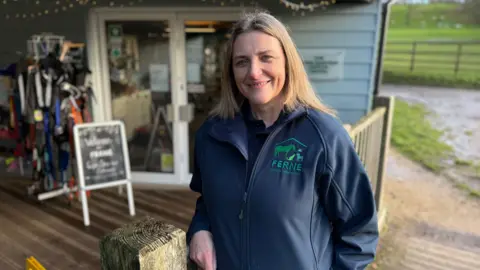 Emma Purnell wearing a dark blue Ferne Animal Sanctuary jacket, and leaning on the fence outside of the charity café in Somerset. A sign saying welcome to Ferne can be seen in the background.