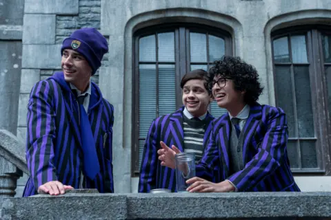 Netflix (L to R) Georgie Farmer as Ajax, Isaac Ordonez as Pugsley Addams, Moosa Mostafa as Eugene in the second season of Wednesday, all three wearing distinctive black and purple blazers. Farmer wears a purple beanie hit with a yellow crest and a purple tie. Ordonez wears a black and white stripey jumper and dark tie and Mostafa wears black-rimmed glasses, a white shirt and dark tie.