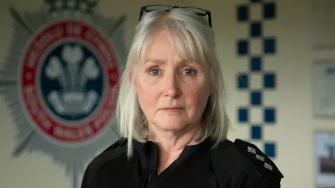 A middle-aged woman, Det Ch Insp Donna Clutterbuck, looks at the camera wearing police uniform standing in front of a South Wales Police logo on a wall