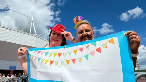 Allison Ellis and Linda Russell holding a banner with images of multi-coloured bunting on it. There are standing in front of a stadium and smiling. There are both wearing colour hats and smiling.