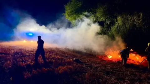 Shefford Fire Station White smoke rising into the air and across a straw field in the night. It is coming from a stack of hay that is glowing red and orange with orange flames. Two firefighters are crouched in front of it and a third is walking across the field. Blue flashing lights from a fire engine can be seen through the smoke.