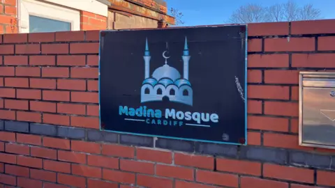 BBC A brick wall with a blue sign reading Madina Mosque
