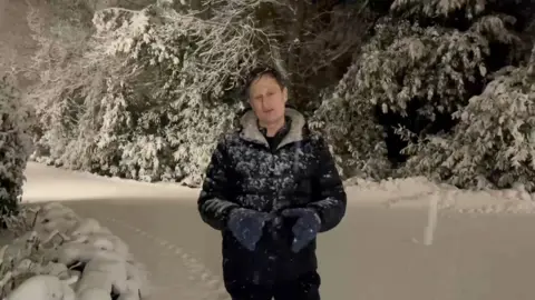 Nicky Schiller wearing a blue winter coat and blue gloves standing in a snow-covered wooded area in Leeds.