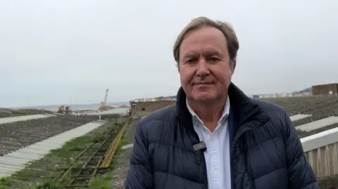 Richard Le Sueur, a man with brown hair, wearing a blue puffa jacket and a white shirt. He is standing in front of a flat, corrugated iron roof.